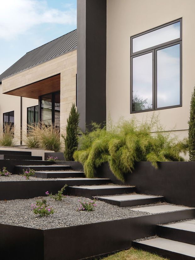 A contemporary residential exterior viewed from a landscaped front garden. The home features pale masonry walls, a dark standing-seam pitched roof, strong black vertical and horizontal elements, large framed windows, and a timber-lined covered entry. Stepped dark retaining walls and concrete pavers form a terraced approach through ornamental grasses and small flowering plants.