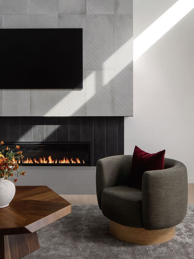 A refined contemporary living room focused on a sculptural upholstered armchair beside a low geometric timber coffee table. A linear fireplace is integrated into a dark vertical-panel feature wall, with a large wall-mounted television above a pale textured geometric wall surface. Strong daylight creates diagonal shadows across the wall, while a deep burgundy cushion adds contrast. The room has a warm, sophisticated and softly dramatic atmosphere.