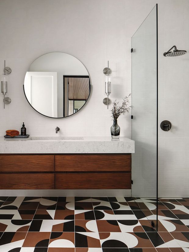 A refined contemporary bathroom featuring a long floating dark timber vanity with a pale stone benchtop, integrated basin and wall-mounted tap. A large round mirror is mounted on a subtly textured light wall beside vertical globe sconces. The shower is enclosed by a frameless glass screen with dark hardware and a round overhead showerhead. The floor has bold geometric tiles in black, white, brown and terracotta tones.