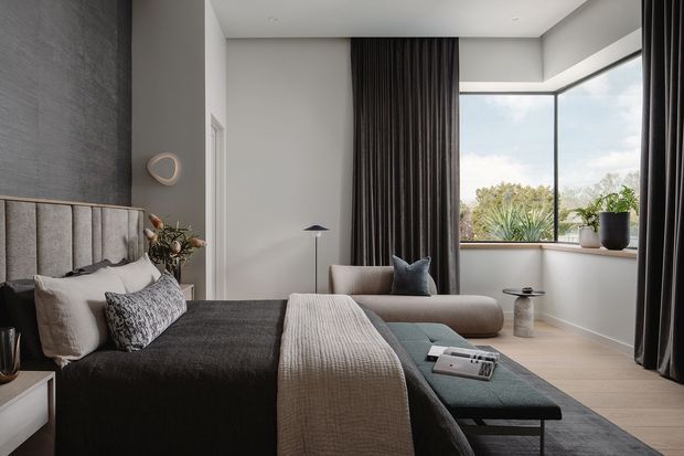 A refined contemporary bedroom with a dark upholstered bed, layered neutral bedding, a softly curved bench, and floor-to-ceiling charcoal curtains. A large corner window brings in natural light and views of greenery, while understated lighting, textured surfaces, and muted grey, beige, and charcoal tones create a calm, luxurious atmosphere.