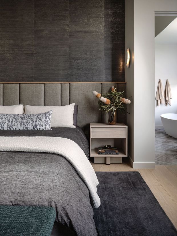 A refined contemporary bedroom in a dark, layered neutral palette. The room features a charcoal textured feature wall, upholstered vertical-panel headboard, grey bedding with a white textured throw, a compact timber bedside table, dark rug, and a sculptural wall light. An open doorway reveals a partially visible bathroom with a freestanding white bathtub and hanging towels.