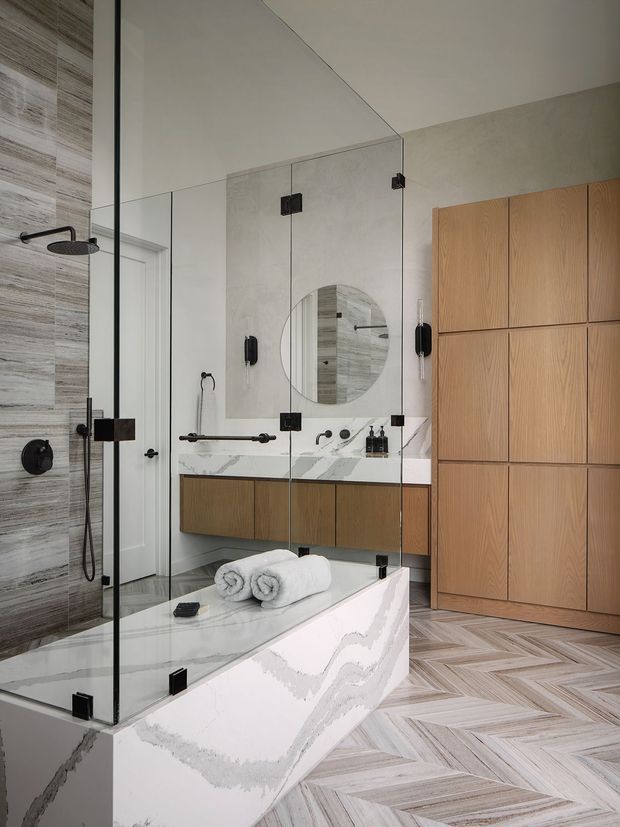A refined contemporary bathroom featuring a freestanding sculptural marble-look bathtub, frameless glass shower enclosure, wall-mounted timber vanity cabinetry, a round mirror, and a tall built-in timber storage wall. Large-format veined stone surfaces and patterned timber-look flooring are paired with matte black fixtures and hardware.