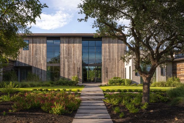 A contemporary single-storey building facade with a broad low-pitched roof and vertically arranged weathered timber cladding. Tall dark-framed glazed openings form a strong central entry composition, with a concrete path leading through a landscaped front garden featuring mature trees, lawn, and planted garden beds.