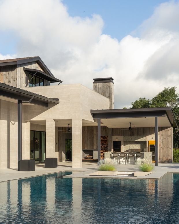 Exterior view of a contemporary residential home arranged around a swimming pool. The house combines pale stone or rendered masonry volumes with weathered timber cladding, dark metal roof edges and framing, large glazed openings, and a covered outdoor kitchen and dining area beside the pool. The composition is bright and refined, with strong indoor-outdoor connection and a partly pitched-roof, partly flat-roof form.
