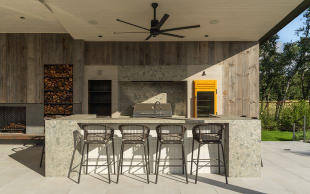 A covered outdoor entertaining area with a built-in kitchen and bar island. The space features a large pale stone-clad counter with four woven bar stools, timber-look wall cladding, a concrete or stone backsplash, integrated grill and shelving, a bright yellow appliance or cabinet, a ceiling fan, and a fireplace at the left. The open side looks onto a garden, creating a contemporary indoor-outdoor setting.