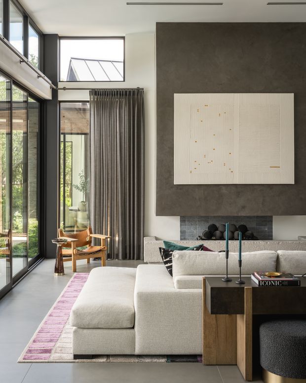 A sophisticated contemporary living room with a large pale sectional sofa arranged around a dark wood coffee table. The room features floor-to-ceiling black-framed glazing, tall grey curtains, a polished concrete floor, and a dramatic dark textured feature wall with a large abstract artwork above a tiled fireplace. Natural light, restrained furnishings, and high ceilings create an airy yet refined atmosphere.