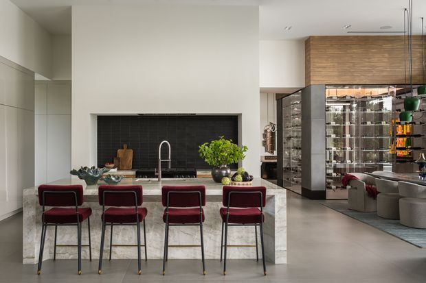 A spacious, open-plan contemporary kitchen and dining area. The kitchen features a large pale stone island with four burgundy upholstered bar stools, white integrated cabinetry, a black tiled backsplash, and a dark cooktop with a sculptural faucet. A warm timber-clad upper wall and extensive glass-fronted storage are visible to the right, along with a dining or living area beyond. The room has a refined, high-end material palette and clean architectural lines.