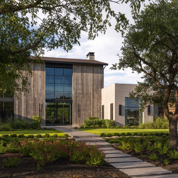Exterior view of a refined contemporary residence set within a landscaped garden. The main volume features weathered vertical timber cladding, a broad dark metal roof, and a tall grid-patterned glazed entry facade. A contrasting pale rendered or concrete wing projects behind it, with large dark-framed windows. A paved stepping path crosses the lawn and planted garden toward the entrance, while mature trees frame the composition.