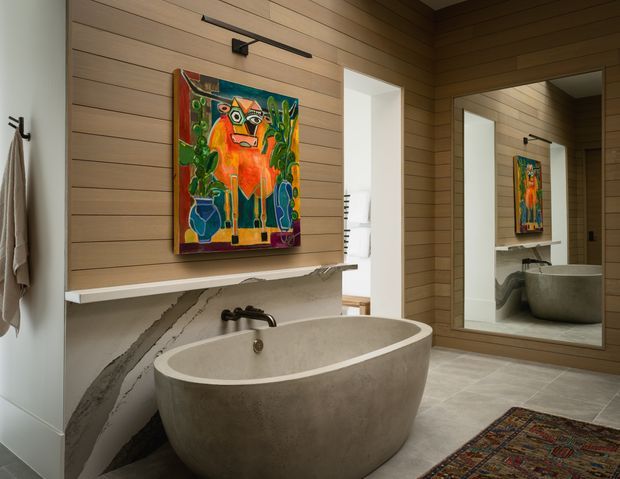 A warm, refined bathroom featuring a freestanding oval stone bathtub, wall-mounted black tapware, a marble-look ledge and splashback, horizontal timber wall panelling, a large framed mirror, and colourful artwork. A partial towel rail and adjacent doorway are visible, with a patterned rug adding texture.
