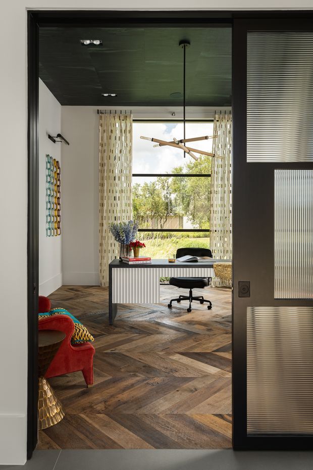 A stylish compact home office viewed through a dark-framed glazed doorway. The room features a sculptural white desk with vertical ribbed detailing, a black task chair, patterned curtains, a large garden-facing window, a dark reflective ceiling, and herringbone timber flooring. A colourful upholstered chair and decorative wall hanging add vivid accents, while the ribbed translucent glass door and black framing create a refined, contemporary feel.