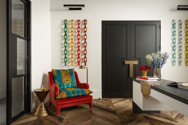 A bold, eclectic entryway with a black double door featuring geometric panel detailing, a black-framed glazed screen door at left, and colourful chain wall art arranged on white walls. A vivid red armchair with patterned blue and yellow upholstery sits beside a sculptural metallic side table. The space has a dark herringbone timber floor, contemporary wall lighting, and a partial white-and-black console or kitchen-like joinery area with decorative objects and flowers.