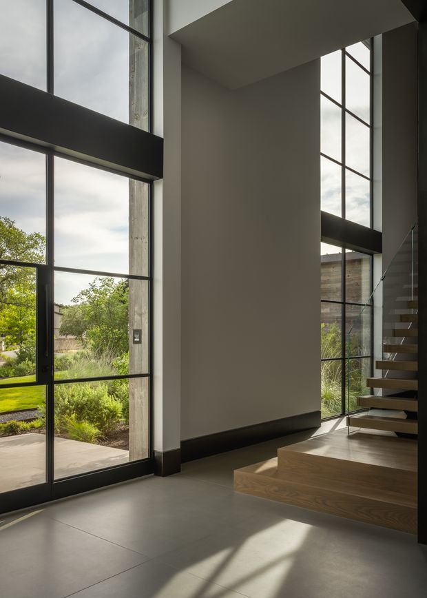 A dramatic double-height residential entry hall with expansive black-framed floor-to-ceiling glazing, a central painted wall volume, and a floating timber staircase with open risers. Concrete-look flooring, dark trim, timber stair detailing, and strong natural light create a refined contemporary interior. Landscaped garden areas are visible through the glazing.