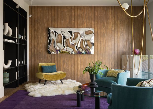 A richly layered living room with a full-height dark timber wall, black built-in display shelving, jewel-toned teal and olive seating, a deep purple rug, and an abstract metallic artwork. A sculptural brass pendant hangs overhead, while a glass coffee table and textured sheepskin add contrast. The room has a polished, eclectic mid-century-inspired character with strong colour and material contrast.
