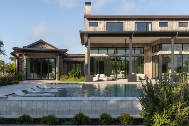 Exterior view of a contemporary multi-level residence with extensive floor-to-ceiling glazing, timber-clad upper walls, a covered outdoor living area, and a tiled swimming pool in the foreground. The building combines a main rectangular volume with a smaller gabled pavilion, set within landscaped gardens.