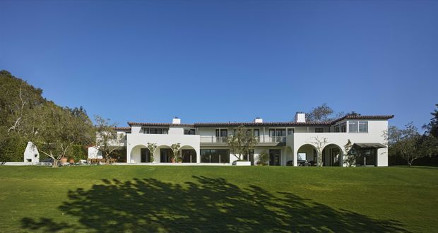 A broad exterior view of a substantial two-storey white stucco residence set behind an expansive manicured lawn. The building has a long, symmetrical composition with repeated rounded arches, dark-framed glazing, projecting balconies, and a low terracotta-tiled roof with several chimneys. Mature trees frame the property, while the clear blue sky and open lawn emphasize its spacious landscaped setting.