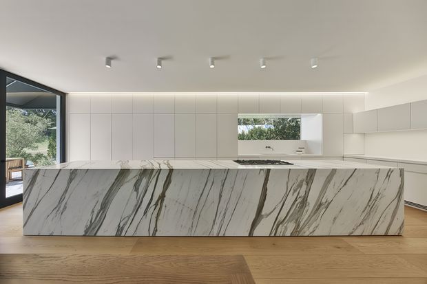 A spacious contemporary kitchen with a long white island featuring dramatic grey and dark veining, floor-to-ceiling pale cabinetry, integrated appliances, recessed linear lighting, and warm timber flooring. Large windows provide views to greenery, while the overall design is clean, minimal, and refined.