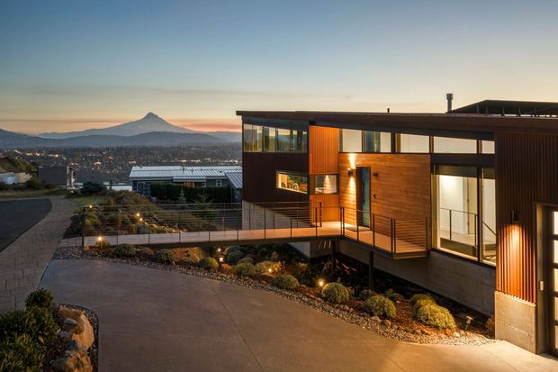 Contemporary multi-level residential home on a sloped suburban site, photographed at dusk. The house features strong horizontal rooflines, extensive glazing, dark metal framing, warm timber wall cladding, elevated terraces and carefully landscaped gravel garden beds. A mountain and suburban settlement are visible in the distance.