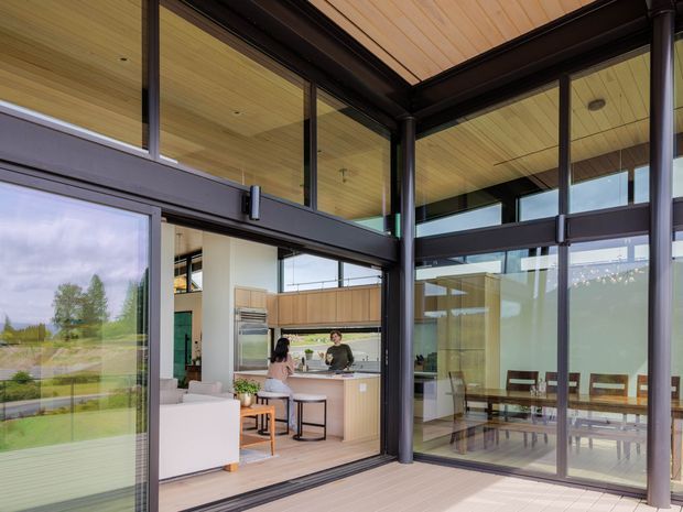 View from a covered outdoor deck or terrace into a contemporary open-plan home through extensive dark-framed glazing. The interior clearly shows a kitchen with island seating, adjacent living area, and a dining table, while timber-lined ceilings and deck surfaces create a warm natural finish. The dark structural framing and floor-to-ceiling windows give the space a strong indoor-outdoor connection.