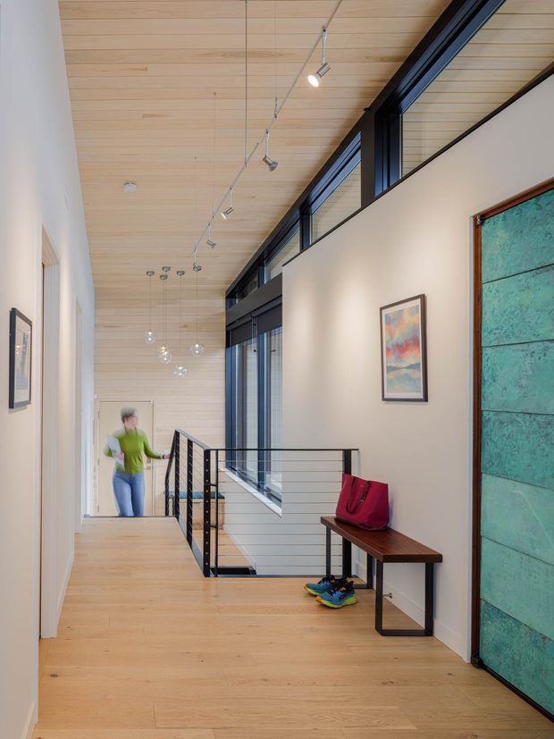 A bright, contemporary interior hallway and stair landing with light timber flooring and a matching timber-lined ceiling. Black-framed clerestory and full-height glazing bring in abundant natural light, while a slim black metal balustrade borders the stair opening. A narrow dark timber console, artwork, pendant lights, track lighting, and a large turquoise-green textured door add visual interest.