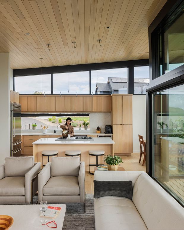 An open-plan contemporary kitchen and living area with extensive black-framed glazing. The kitchen features light timber cabinetry, a large white island with integrated cooktop and seating, stainless-steel appliances, and a timber-lined ceiling. In the foreground are neutral upholstered armchairs and a curved sofa around a coffee table, with a dining area visible behind the kitchen. The space is bright, warm, and connected to the outdoors through large windows and glass doors.