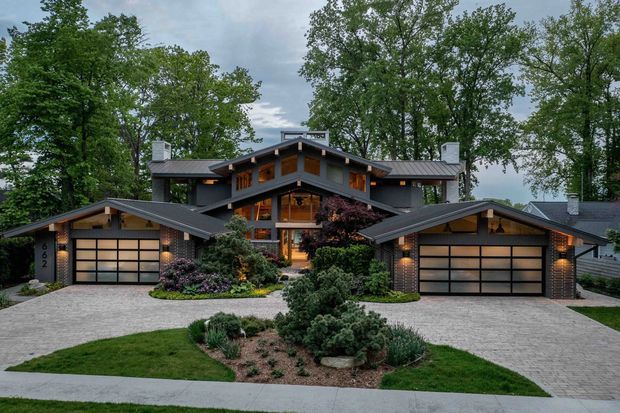 Front exterior view of a large, symmetrical multi-level residence set among mature trees and landscaped gardens. The dark grey pitched roof, projecting gabled wings, exposed timber detailing, warm wood-framed glazing, stone or brick-clad columns, and twin integrated garage wings create a substantial lodge-like composition. A paved driveway and central garden bed lead to the recessed entry.