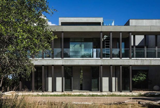 A two-storey contemporary residential facade with a strong horizontal composition, flat rooflines, exposed concrete bands, dark vertical columns, extensive glazing, and upper-level glass balustrades. The building is set among mature landscaping and photographed in bright daylight against a clear blue sky.