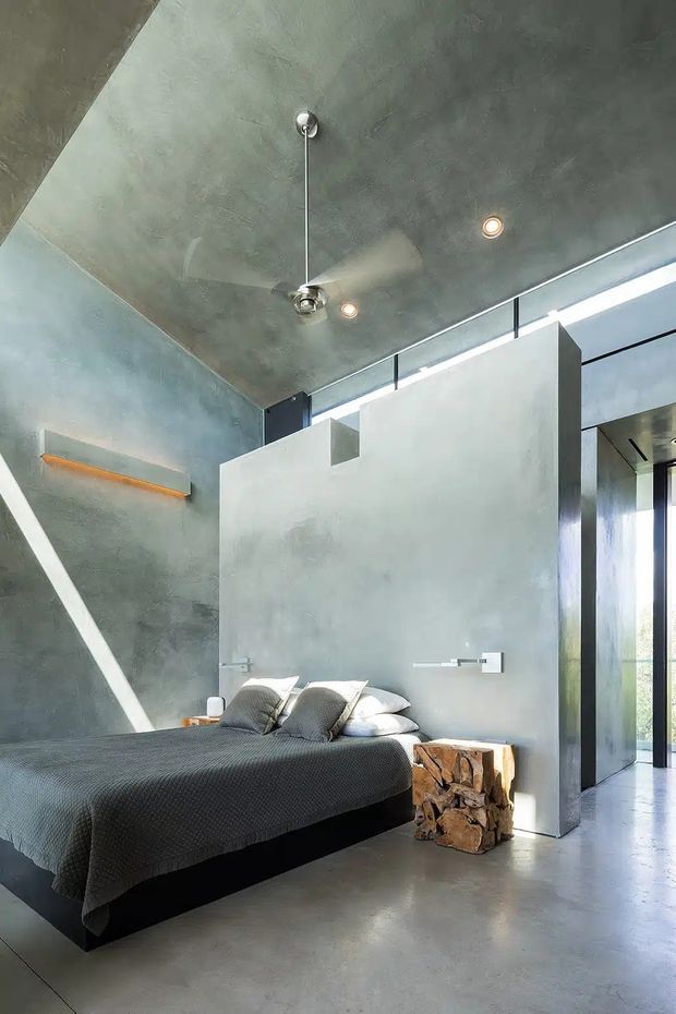 A dramatic contemporary bedroom with a low platform bed dressed in charcoal bedding, set against raw grey concrete walls and ceiling. A ceiling-mounted fan, recessed spotlights, narrow clerestory glazing, and large black-framed openings create an industrial, minimalist atmosphere. A sculptural rough-hewn timber stool sits beside the bed, while strong sunlight enters from the side.