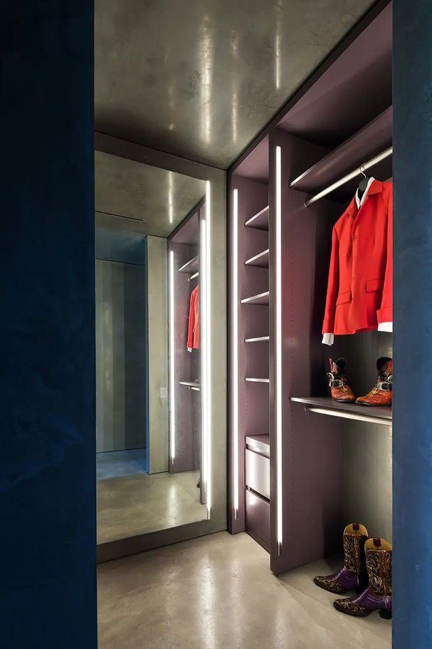 A narrow, dramatically lit walk-in wardrobe with dark purple built-in shelving, hanging rails, drawers, integrated vertical LED lighting, and a full-height mirror. Clothing and boots are displayed within the joinery. The space has a polished concrete floor and ceiling, with deep blue surrounding walls and a dark, moody palette.