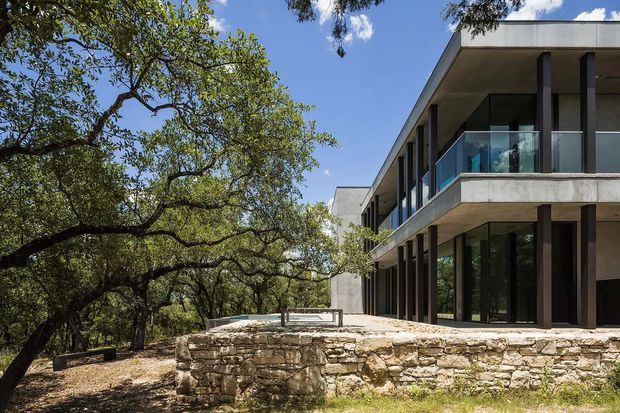 Exterior view of a contemporary two-storey residence set among mature trees. The building has strong horizontal lines, a flat roof, deep overhangs, extensive dark-framed glazing, glass balcony balustrades, and pale rendered or concrete surfaces. A rustic natural-stone retaining wall and terrace connect the home to the wooded landscape.