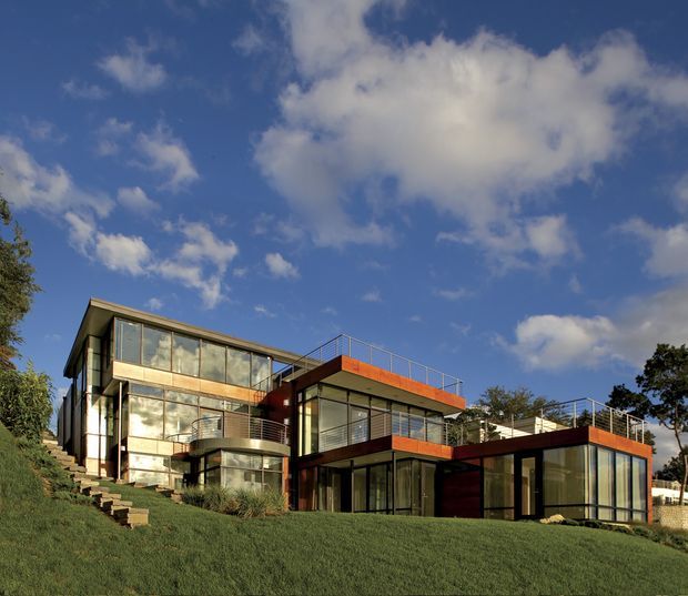 Exterior view of a multi-level contemporary house set into a sloping grassy site. The building has extensive floor-to-ceiling glazing, dark structural framing, warm orange-brown horizontal roof and fascia elements, projecting box forms, balconies or roof terraces with slim metal railings, and a curved glazed corner volume. Mature trees and a blue sky surround the property.