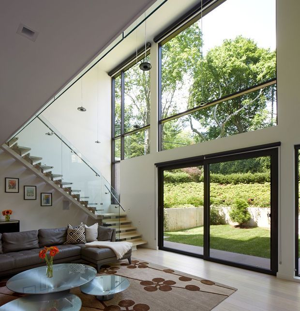 A bright contemporary living room with a double-height glazed wall overlooking a landscaped garden. The space features a large dark grey sectional sofa, glass coffee tables, patterned area rug, floating timber stair with clear glass balustrade, pendant lights, and extensive black-framed windows and sliding doors.