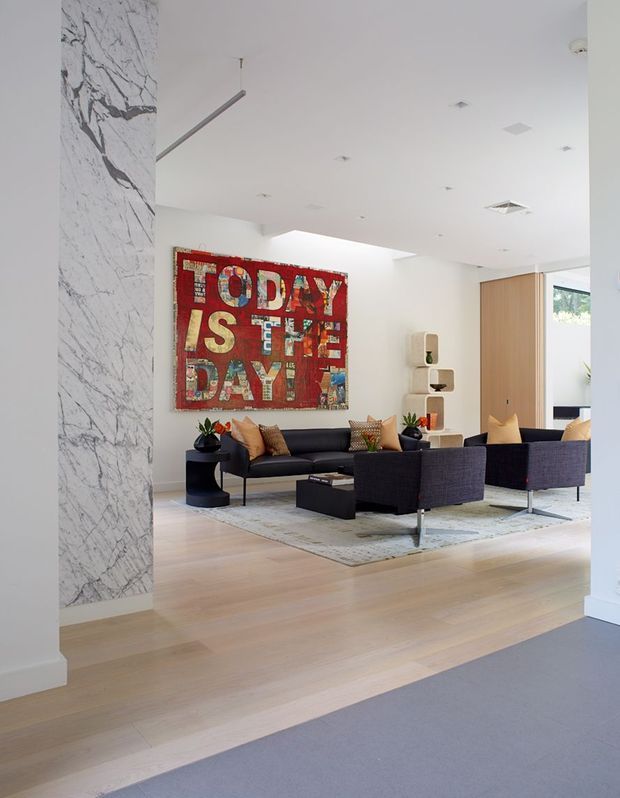 Bright contemporary living room viewed from an adjoining carpeted area, featuring pale timber flooring, a large white-and-grey veined marble-clad column, white walls and ceiling, dark charcoal sofas and armchairs, a light area rug, and a bold red typographic artwork. Timber panelled sliding or pivot doors and minimalist recessed lighting are visible at the rear.