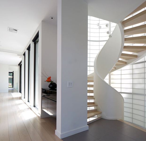 Bright contemporary interior hallway featuring a sculptural white spiral staircase with light timber open treads, a curved translucent glass-block and glazed wall, and a tall multi-light pendant. Pale timber flooring, white walls, streamlined black-framed glazing, and minimal built-in joinery create an airy, refined space.