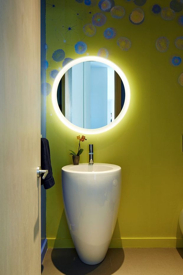 A compact, vividly styled bathroom viewed through an open doorway. The room has a lime-green feature wall decorated with blue and gold circular motifs, a sculptural white pedestal basin with a chrome tap, a round backlit mirror, dark tiled flooring, and a small orchid on the basin. A towel hangs beside the door.