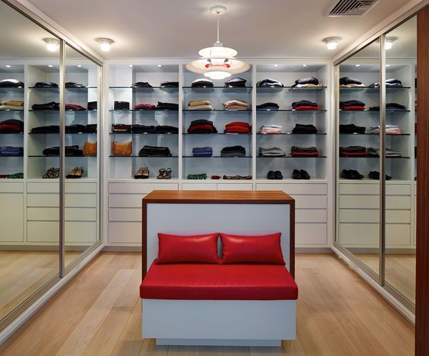 A polished walk-in wardrobe with extensive white built-in shelving and drawers displaying folded clothing, handbags, and shoes. Mirrored sliding doors line both sides of the room and reflect the storage, while a central upholstered red bench with cushions provides seating. Light timber flooring, recessed ceiling lights, and a contemporary pendant create a bright, refined dressing-room interior.