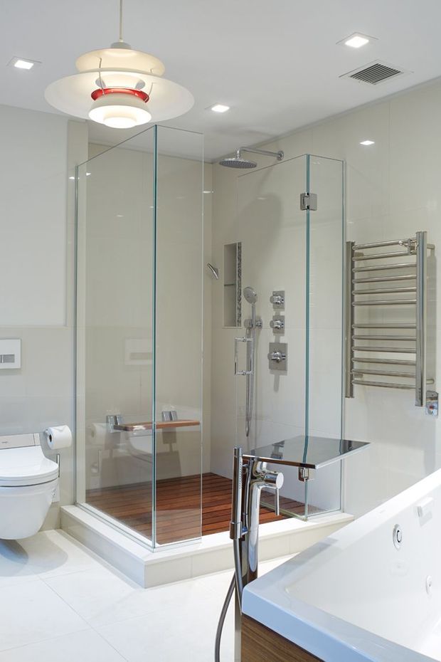 Bright contemporary bathroom featuring a large frameless glass walk-in shower with multiple chrome controls, overhead rainfall shower, handheld shower, and timber-look shower floor. A freestanding white bathtub, wall-mounted chrome towel warmer, toilet, recessed lighting, and sculptural pendant light complete the space.