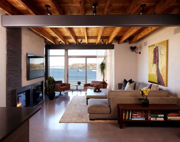A warm, waterfront-facing living room with expansive glazing, a contemporary sectional sofa, fireplace and television wall, and a striking exposed timber-beam ceiling. The room opens toward a calm blue body of water, with polished concrete flooring, a large neutral rug, and rich wood and earthy furnishings.