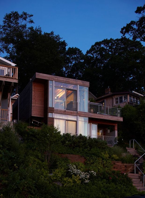 A dusk exterior photograph of a compact, multi-level contemporary house set into a densely planted, steeply sloping site. The building features dark timber cladding, broad glass openings, projecting balconies with slim horizontal metal railings, and stepped exterior stairs rising through landscaped terraces. Mature trees frame the home against a deep blue sky.