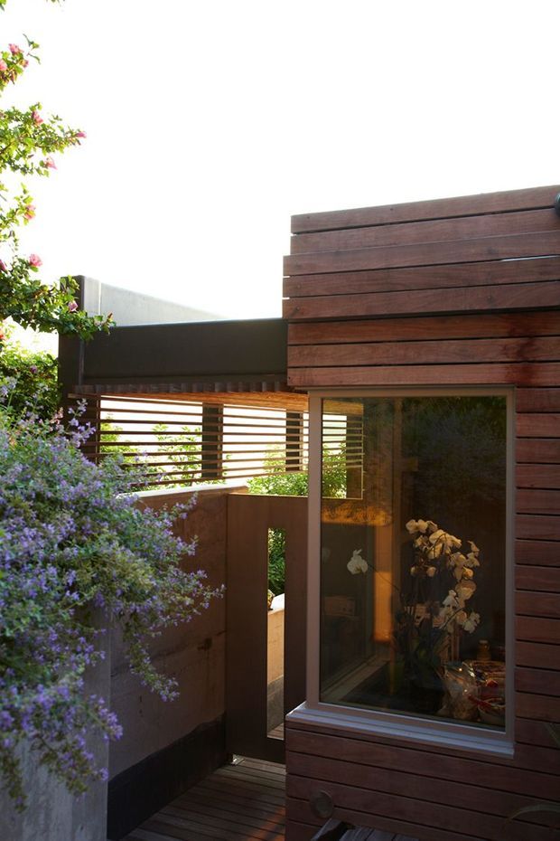 A narrow outdoor passage beside a modern timber-clad building. Dark horizontal wood boards form the facade, with a large window and glazed door opening onto a small timber deck. A slatted privacy screen and dark metal fencing define the courtyard-like walkway, while dense flowering planting softens the edges. The image is brightly lit and focuses on the exterior materials, screening, glazing, and garden setting.
