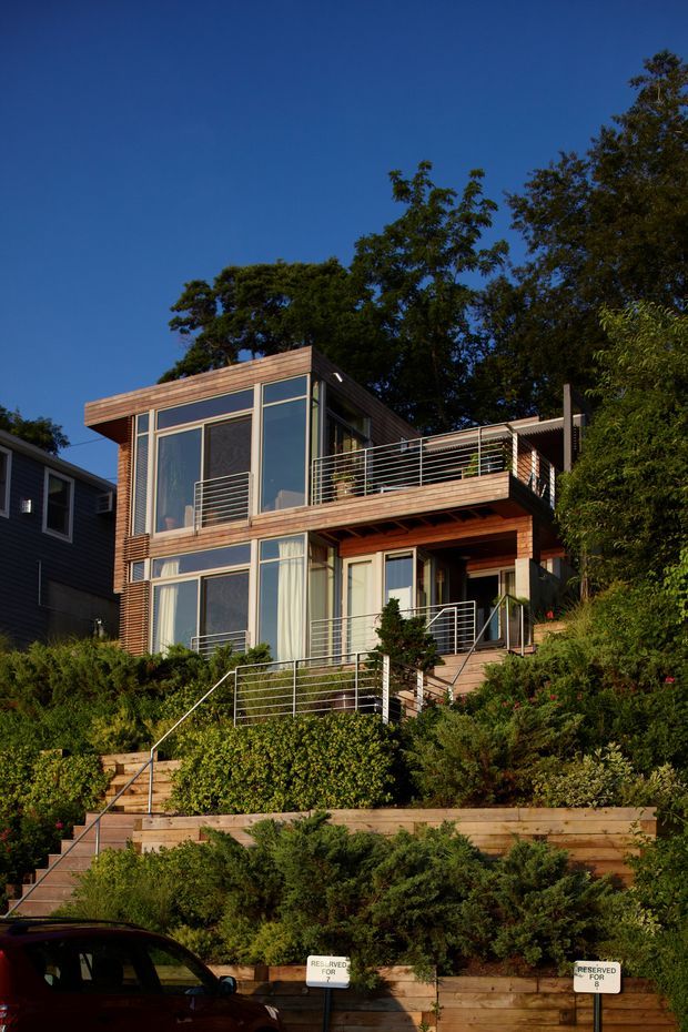 Exterior view of a compact multi-level contemporary house set on a steep, heavily landscaped site. The home features extensive floor-to-ceiling glazing, horizontal timber cladding and trim, projecting balconies and terraces with slim metal railings, and stepped timber retaining walls with stairs leading up from the street.