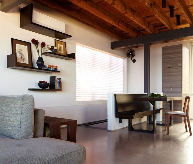 A warm, compact open-plan living and dining area with an exposed timber ceiling, polished concrete floor, white walls, floating dark shelving, and dark structural steel beams. The room includes a grey upholstered sofa, compact timber side table, wall-mounted air conditioner, built-in dining/work bench, timber dining table and chairs, and layered window coverings. The combination of exposed wood and steel creates a contemporary industrial character with warm natural detailing.