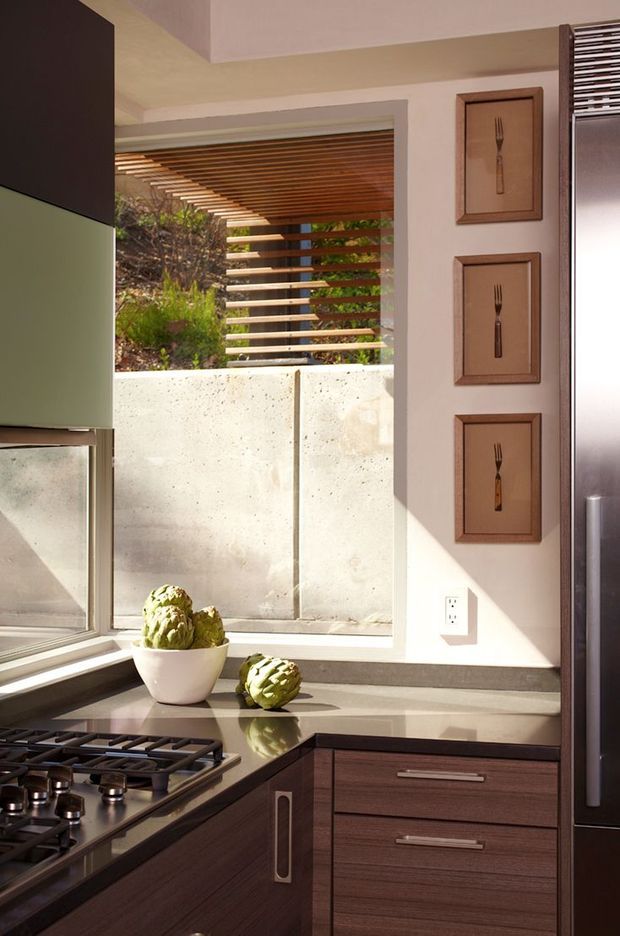 A warmly lit contemporary kitchen corner with dark wood-grain cabinetry, a dark stone benchtop, stainless-steel appliances and a gas cooktop. A large window opens toward a landscaped courtyard, shaded by a timber louvre pergola. Three vertically arranged framed fork artworks add a decorative accent to the pale wall.