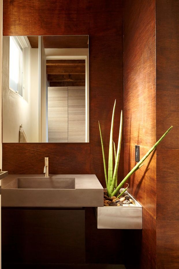 Compact contemporary bathroom with a wall-mounted rectangular concrete or stone vanity, integrated basin, slim metal faucet, frameless mirror, and a built-in planter filled with stones and a tall snake plant. Rich dark timber wall panelling and ceiling surfaces create a warm, dramatic interior, with a small window providing natural light.