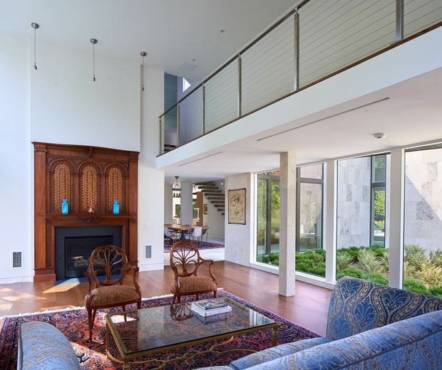 A bright, open-plan living room with a double-height ceiling and mezzanine gallery. The room features a highly ornate dark timber fireplace surround, patterned Persian-style rug, carved wood armchairs, a glass-topped coffee table, and a blue patterned sofa. Floor-to-ceiling glazing opens views to a landscaped garden, while a stairway and dining area are visible beyond.