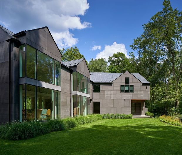 Exterior view of a contemporary multi-wing residential house set within a landscaped lawn and mature trees. The building features dark grey and taupe cladding, steep gabled roof forms, extensive floor-to-ceiling glazing, projecting glass-fronted volumes, and restrained rectangular windows.
