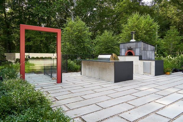 A contemporary outdoor entertaining area set within a lush wooded garden. Large pale rectangular paving slabs form the terrace, with a built-in outdoor kitchen and dark grey masonry pizza oven. A bold red rectangular portal frames the entrance, while glass and dark metal screening define the edge of the space.