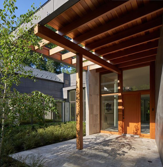 Exterior architectural view of a contemporary entry courtyard or covered terrace. A deep timber pergola with exposed beams and rafters shelters a paved approach to a timber front door and tall glazed openings. The facade combines pale stone or concrete surfaces with dark cladding, while landscaped planting and a neighbouring roof are visible beyond.