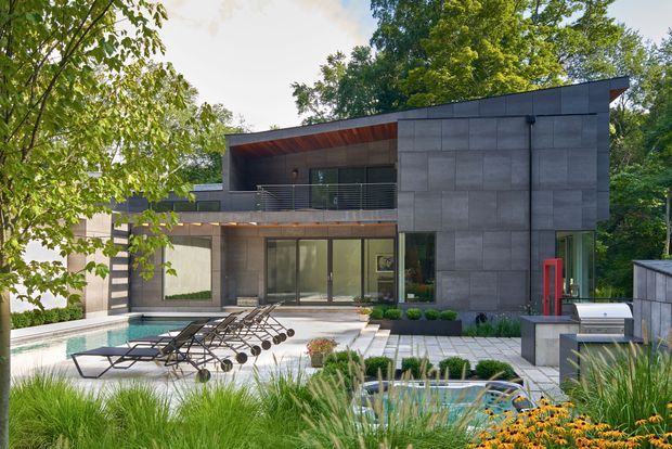 A contemporary two-storey house set among mature trees, viewed from a landscaped pool area. The home has a strong geometric form, dark grey large-format facade panels, extensive floor-to-ceiling glazing, a deep upper-level balcony with horizontal metal balustrading, and warm timber soffit lining. A swimming pool, paved terrace, loungers, built-in outdoor kitchen or grill area, and ornamental planting occupy the foreground.