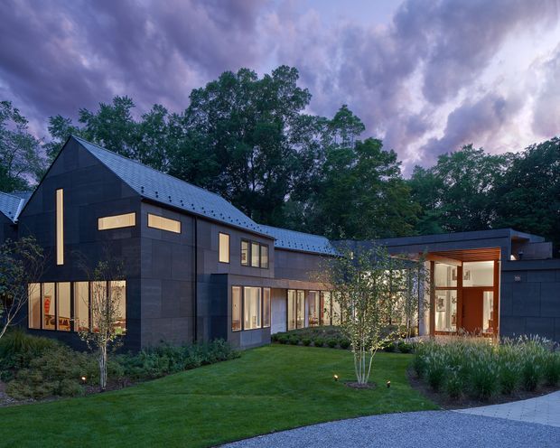 Exterior architectural photograph of a contemporary dark-clad residence set within a landscaped garden at dusk. The composition shows a steep gabled wing with narrow vertical and horizontal windows, connected to a lower flat-roofed volume with extensive glazing and a sheltered timber-lined entry. Mature trees, lawn, planting beds, and a paved foreground surround the house beneath a dramatic cloudy sky.