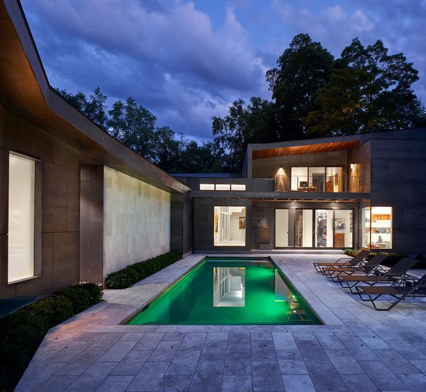 Twilight view of a sophisticated modern courtyard pool area enclosed by dark rectilinear architecture. A narrow illuminated swimming pool is surrounded by large-format pale paving, built-in landscaping, and rows of dark loungers. Floor-to-ceiling glazing opens onto the courtyard, while warm recessed and wall lighting contrasts with the deep blue sky and surrounding mature trees.